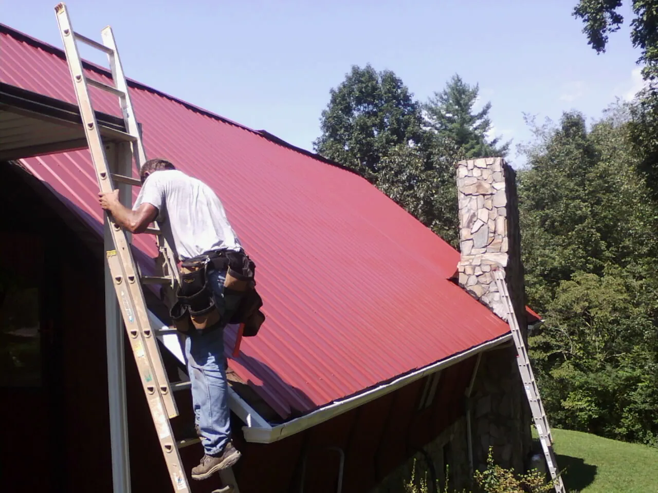Roofer on a ladder beside a red metal roof and stone chimney in Big Indian